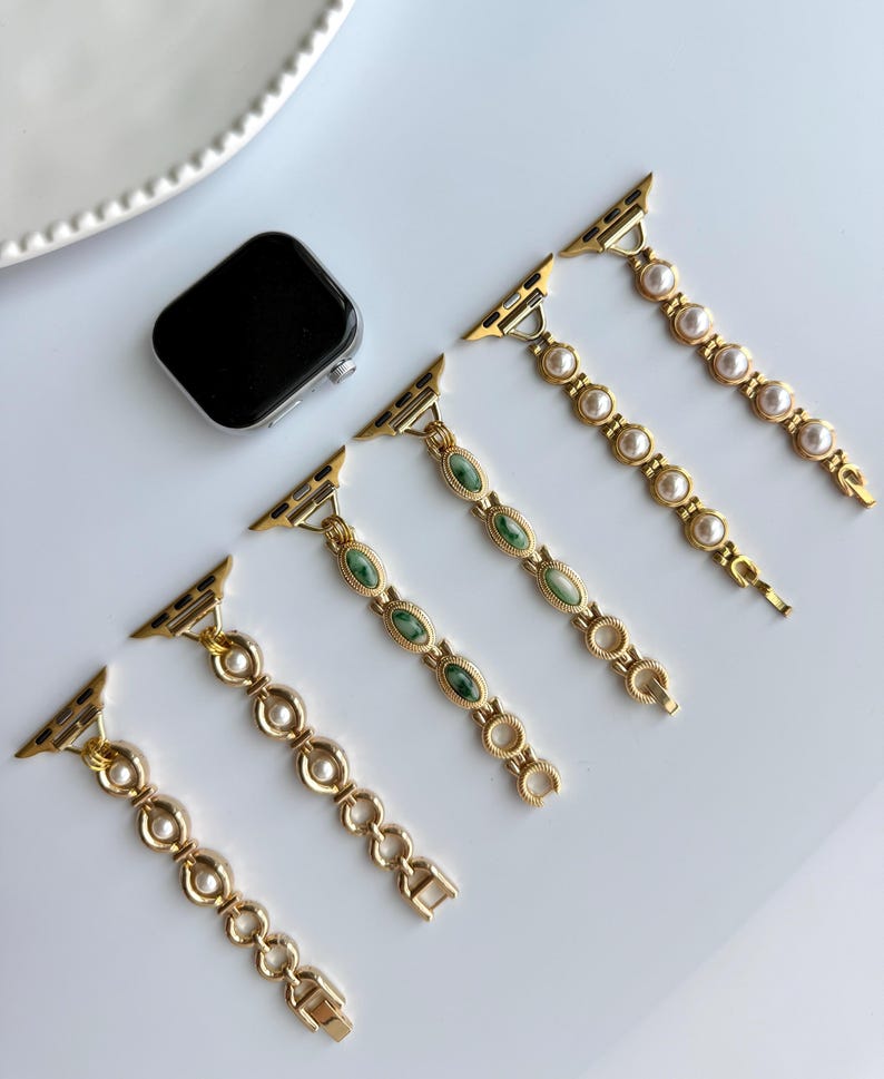 May include: Gold-toned watch bands with pearl and green stone accents. The bands are displayed with a black smartwatch on a white surface. The bands feature a variety of designs, including oval green stones and round pearls.