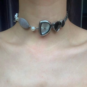 Puede incluir: Un collar gargantilla plateado con una esfera de reloj triangular, una piedra ovalada gris y detalles negros en forma de corazón. El collar también incluye una pequeña perla y una cadena plateada.