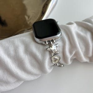 May include: A silver-toned smartwatch with a black screen and a metallic link watch band. The band features decorative charms including a star shape encrusted with clear crystals and a small heart-shaped charm with a white enamel-like finish.