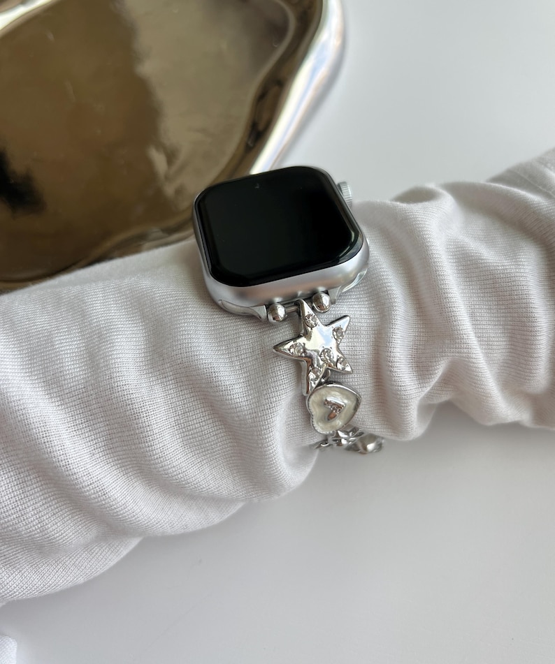 May include: A silver-toned smartwatch with a black screen and a metallic link watch band. The band features decorative charms including a star shape encrusted with clear crystals and a small heart-shaped charm with a white enamel-like finish.