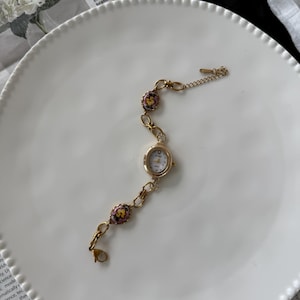 May include: A gold-toned wristwatch with a small, round white face and delicate floral accents. The watch features a chain-link band with decorative floral beads in pink, yellow, and green. The watch is set on a white plate.