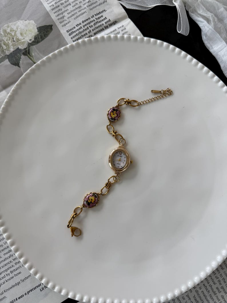 May include: A gold-toned wristwatch with a small, round white face and delicate floral accents. The watch features a chain-link band with decorative floral beads in pink, yellow, and green. The watch is set on a white plate.