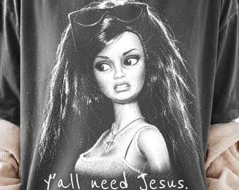 Może przedstawiać: Ciemnoszary t-shirt z czarno-białą grafiką przedstawiającą lalkę z długimi ciemnymi włosami, noszącą okulary przeciwsłoneczne na głowie i naszyjnik z krzyżykiem. Poniżej ilustracji znajduje się biały napis "y'all need Jesus.".
