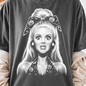 May include: A charcoal gray t-shirt featuring a white graphic illustration of a person with a high bun hairstyle, wearing a large bow and a halter top decorated with pumpkin and spiderweb patterns. The person has a wide-eyed, surprised expression.