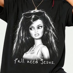 Może przedstawiać: Czarny t-shirt z białą grafiką przedstawiającą lalkę z długimi włosami, okularami przeciwsłonecznymi na głowie i naszyjnikiem z krzyżykiem. Napis "Y'all need Jesus." jest nadrukowany na biało pod ilustracją lalki na koszulce.
