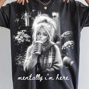 Puede incluir: Una camiseta de manga corta gris carb&oacute;n con un estampado gr&aacute;fico en blanco y negro de una persona con un mo&ntilde;o desordenado, bufanda y sosteniendo una taza de caf&eacute;. El fondo muestra &aacute;rboles y calabazas. El texto "mentally i'm here" est&aacute; impreso en blanco.