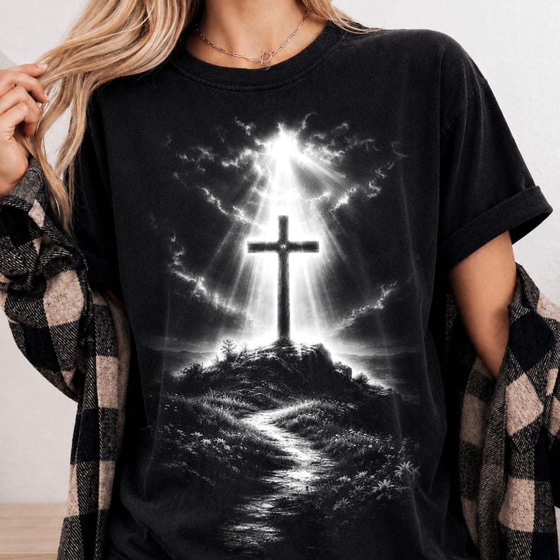 May include: A black short-sleeve t-shirt featuring a graphic print of a cross standing on a hill. The design includes a bright light radiating from behind the cross, illuminating the sky and a path leading up the hill in a monochromatic style.