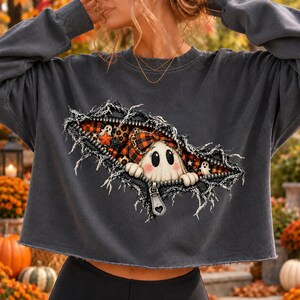 May include: A charcoal gray long-sleeve sweatshirt featuring a graphic of a zipper opening to reveal a ghost wearing a plaid bow. The design includes orange and black plaid patterns, leopard print accents, and small ghost figures on a dark background.