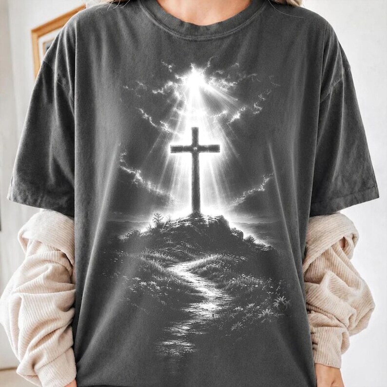 May include: A charcoal gray short-sleeve t-shirt featuring a graphic print of a cross standing atop a hill. The design includes a bright light emanating from behind the cross, illuminating the sky and a path leading up the grassy hillside.