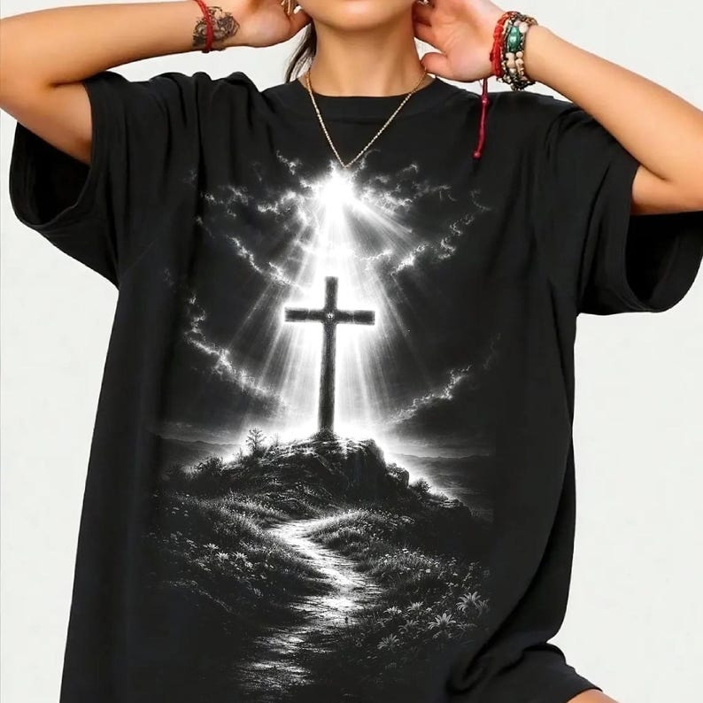 May include: A black short-sleeve t-shirt featuring a graphic print of a cross standing atop a hill. The cross is illuminated by bright white light rays emanating from behind it against a dark, cloudy sky. A path leads up the hill through grass.