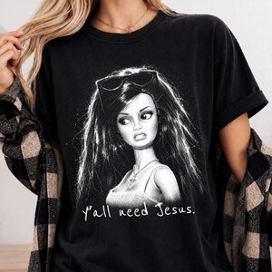 Może przedstawiać: Czarny t-shirt z kr&oacute;tkim rękawem z białą grafiką przedstawiającą lalkę z długimi włosami i okularami przeciwsłonecznymi na głowie. Lalka ma na szyi naszyjnik z krzyżykiem. Poniżej grafiki znajduje się nadruk z tekstem "y'all need Jesus.".