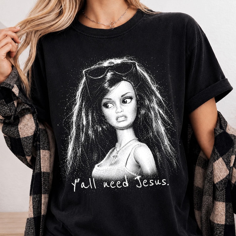 Może przedstawiać: Czarny t-shirt z kr&oacute;tkim rękawem z białą grafiką przedstawiającą lalkę z długimi włosami i okularami przeciwsłonecznymi na głowie. Lalka ma na szyi naszyjnik z krzyżykiem. Poniżej grafiki znajduje się nadruk z tekstem "y'all need Jesus.".