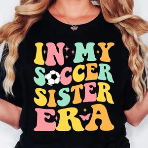 May include: Black t-shirt with the text "IN MY SOCCER SISTER ERA" in colorful, retro-style lettering. A soccer ball graphic is in the center. The design includes stars and a butterfly.