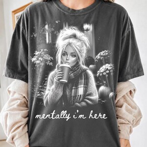 Puede incluir: Una camiseta gris carb&oacute;n con un gr&aacute;fico en blanco y negro de una persona con el cabello en un mo&ntilde;o desordenado, usando una bufanda y sosteniendo una taza de caf&eacute;. El fondo muestra calabazas y flores. El texto "mentally i'm here" est&aacute; impreso.