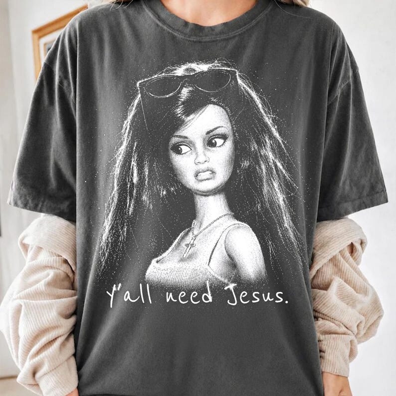Może przedstawiać: Ciemnoszary t-shirt z czarno-białą grafiką przedstawiającą lalkę z długimi ciemnymi włosami, noszącą okulary przeciwsłoneczne na głowie i naszyjnik z krzyżykiem. Poniżej ilustracji znajduje się biały napis "y'all need Jesus.".