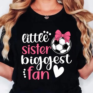 May include: Black t-shirt with the text "little sister biggest fan" in white and pink. A soccer ball with a pink bow is featured above the text. A white heart is below the text. The shirt is a casual style.