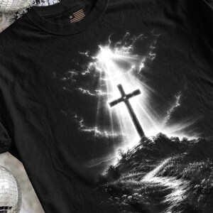 May include: A black t-shirt featuring a graphic print of a Christian cross standing atop a rocky hill. The scene is rendered in black and white with a bright, radiant light shining down from the sky behind the cross, illuminating the clouds.