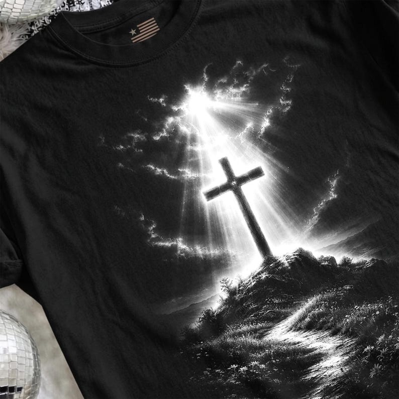 May include: A black t-shirt featuring a graphic print of a Christian cross standing atop a rocky hill. The scene is rendered in black and white with a bright, radiant light shining down from the sky behind the cross, illuminating the clouds.