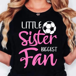 May include: Black t-shirt with the text "LITTLE Sister BIGGEST Fan" in white and pink. A soccer ball graphic is above the word "Sister". The shirt is a crew neck.