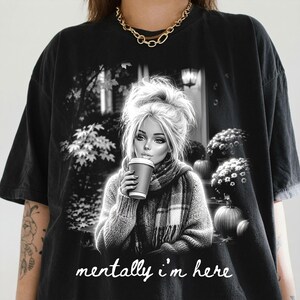 Puede incluir: Una camiseta negra de manga corta con un estampado gr&aacute;fico de una persona con el pelo recogido en un mo&ntilde;o, usando una bufanda y sosteniendo una taza de caf&eacute;. La escena incluye calabazas y follaje. El texto "mentally i'm here" est&aacute; impreso en letra cursiva blanca debajo del gr&aacute;fico.