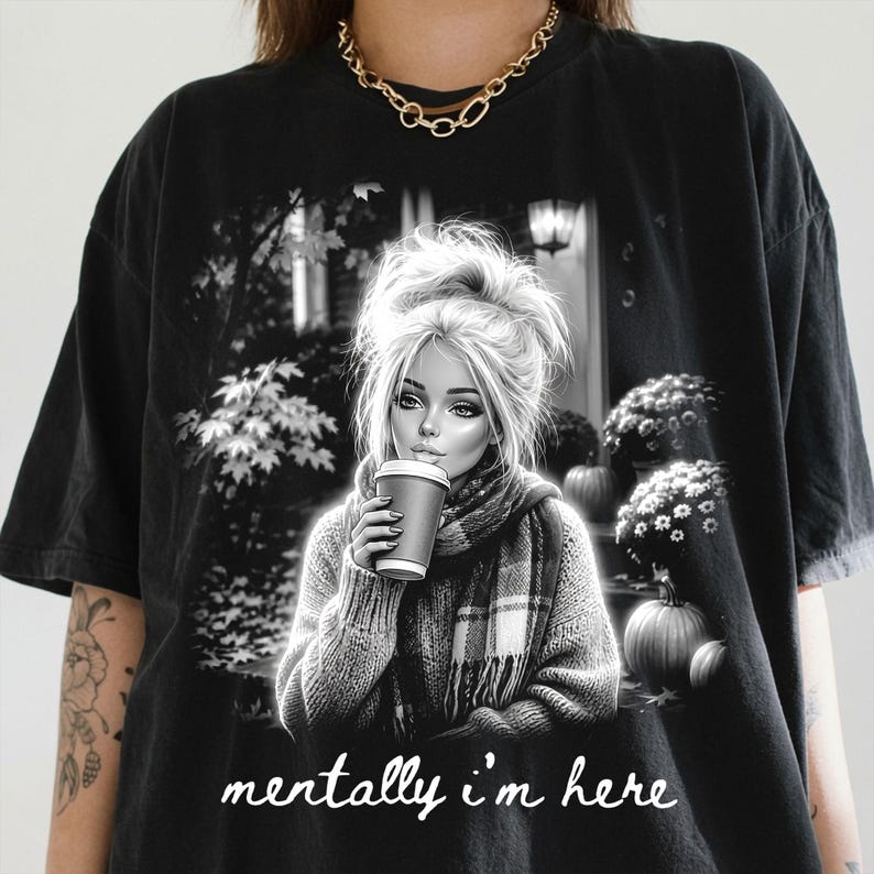 Puede incluir: Una camiseta negra de manga corta con un estampado gr&aacute;fico de una persona con el pelo recogido en un mo&ntilde;o, usando una bufanda y sosteniendo una taza de caf&eacute;. La escena incluye calabazas y follaje. El texto "mentally i'm here" est&aacute; impreso en letra cursiva blanca debajo del gr&aacute;fico.