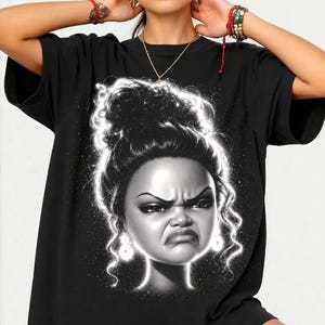 May include: A black short-sleeved t-shirt featuring a large, stylized graphic print of a person with a high bun hairstyle and a grumpy facial expression. The illustration is rendered in black and white with a glowing, ethereal white outline.