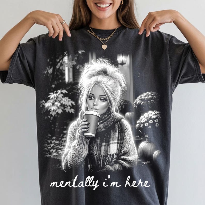 Puede incluir: Una camiseta de manga corta gris carb&oacute;n con un estampado gr&aacute;fico en blanco y negro de una persona con un mo&ntilde;o desordenado, bufanda y sosteniendo una taza de caf&eacute;. El fondo muestra &aacute;rboles y calabazas. El texto "mentally i'm here" est&aacute; impreso en blanco.