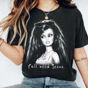 Może przedstawiać: Czarny t-shirt z kr&oacute;tkim rękawem z białą grafiką przedstawiającą lalkę z długimi ciemnymi włosami, okularami przeciwsłonecznymi na głowie i naszyjnikiem z krzyżykiem. Poniżej ilustracji nadrukowano biały napis Y'all need Jesus.