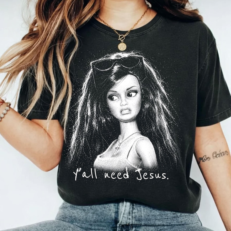 Może przedstawiać: Czarny t-shirt z kr&oacute;tkim rękawem z białą grafiką przedstawiającą lalkę z długimi ciemnymi włosami, okularami przeciwsłonecznymi na głowie i naszyjnikiem z krzyżykiem. Poniżej ilustracji nadrukowano biały napis Y'all need Jesus.