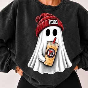 May include: A charcoal black crewneck sweatshirt featuring a graphic of a white ghost wearing a red knit beanie with the text BOO on the front. The ghost is holding a plastic cup of iced coffee with a red straw and a Seven Brew logo on the side.