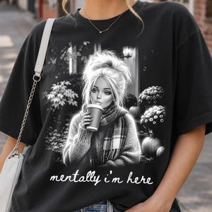 Puede incluir: Una camiseta negra de manga corta con un estampado gr&aacute;fico de una persona con el pelo revuelto, bufanda y una taza de caf&eacute;. La escena incluye calabazas y follaje. El texto "mentally i'm here" est&aacute; impreso en letra cursiva blanca bajo el gr&aacute;fico.