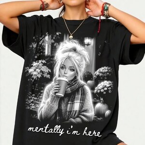 Puede incluir: Una camiseta negra con un estampado gr&aacute;fico de una persona con el pelo revuelto, bufanda y una taza de caf&eacute;. El fondo muestra calabazas y &aacute;rboles. El texto "mentally i'm here" est&aacute; impreso en la parte inferior en letra cursiva de color blanco.