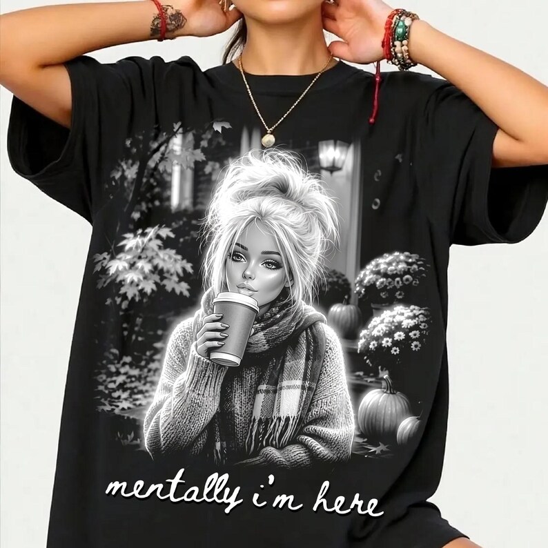 Puede incluir: Una camiseta negra con un estampado gr&aacute;fico de una persona con el pelo revuelto, bufanda y una taza de caf&eacute;. El fondo muestra calabazas y &aacute;rboles. El texto "mentally i'm here" est&aacute; impreso en la parte inferior en letra cursiva de color blanco.