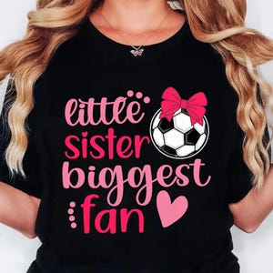 May include: Black t-shirt with the text "little sister biggest fan" in pink script. A soccer ball with a pink bow is above the text, and a pink heart is below. The shirt is a casual style.