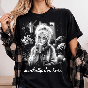 Puede incluir: Una camiseta negra con un gr&aacute;fico en blanco y negro de una persona con mo&ntilde;o, bufanda y una taza de caf&eacute;. La escena incluye calabazas y flores. El texto "mentally i'm here" est&aacute; impreso en color blanco con una tipograf&iacute;a cursiva.