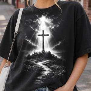 May include: A black short-sleeve t-shirt featuring a graphic print of a cross standing atop a hill. The design depicts a bright light radiating from behind the cross, illuminating a path leading up the hill through a dark, cloudy sky.