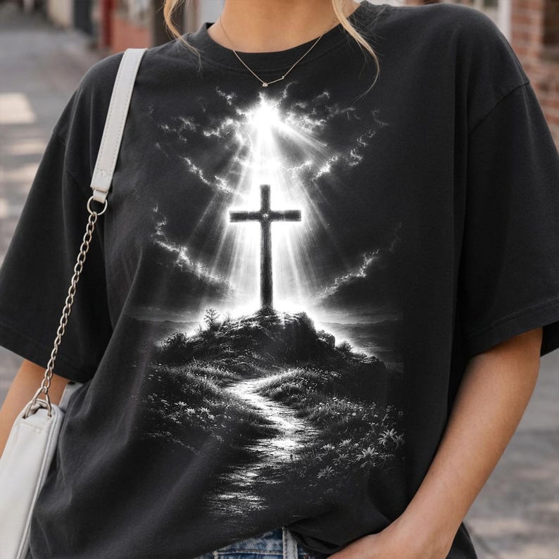 May include: A black short-sleeve t-shirt featuring a graphic print of a cross standing atop a hill. The design depicts a bright light radiating from behind the cross, illuminating a path leading up the hill through a dark, cloudy sky.