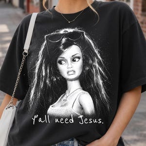 Może przedstawiać: Czarny t-shirt z kr&oacute;tkim rękawem z białą grafiką przedstawiającą lalkę z długimi włosami i okularami przeciwsłonecznymi na głowie. Lalka ma na sobie naszyjnik z krzyżykiem i podkoszulek. Poniżej znajduje się biały napis "y'all need Jesus.".