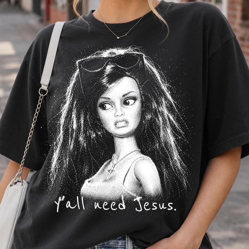 Może przedstawiać: Czarny t-shirt z kr&oacute;tkim rękawem z białą grafiką przedstawiającą lalkę z długimi włosami i okularami przeciwsłonecznymi na głowie. Lalka ma na sobie naszyjnik z krzyżykiem i podkoszulek. Poniżej znajduje się biały napis "y'all need Jesus.".