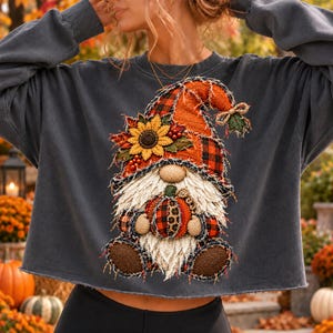 May include: A charcoal gray long-sleeve sweatshirt featuring a graphic of a gnome wearing a plaid hat adorned with a sunflower. The gnome has a long white beard and is holding a small pumpkin with a leopard print pattern on its surface.