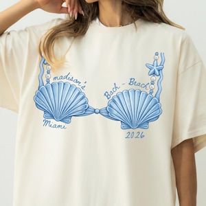 May include: Ivory-colored t-shirt featuring a blue seashell bra design with the text "madison's Bach - Beach Miami 2026". The graphic includes a starfish and pearl accents. The shirt is a relaxed fit.