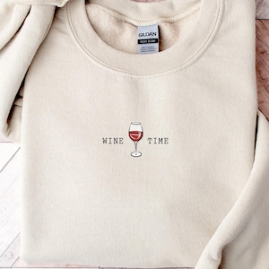 May include: Sand-colored sweatshirt with the words "WINE TIME" and a wine glass graphic embroidered on the front. The long-sleeved top is made of a soft material and is folded on a wooden surface.