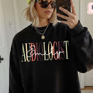 May include: Black sweatshirt with the word "AUDIOLOGIST" in yellow, red, and white lettering, with the name "Emily" in a cursive font. The sweatshirt is a crew neck style.