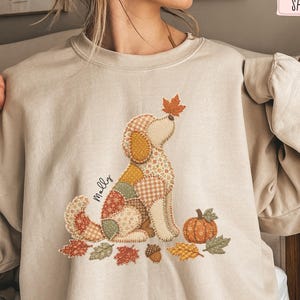 Puede incluir: Una sudadera de cuello redondo color arena con un gráfico de un perro hecho de varios patrones de patchwork en tonos naranja, amarillo y marrón. El perro está rodeado de hojas de otoño, una calabaza y una bellota. El nombre Molly está impreso.