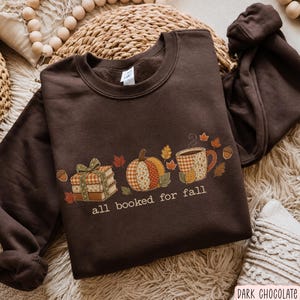 Puede incluir: Una sudadera de cuello redondo color marrón chocolate oscuro con un estampado gráfico de una pila de libros atados con una cinta, una calabaza estampada y una taza humeante, con el texto "all booked for fall" impreso en blanco debajo.