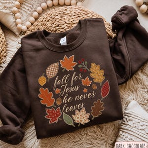 Puede incluir: Una sudadera de cuello redondo en color marrón chocolate oscuro con un gráfico central de una corona hecha de hojas de otoño estampadas, bellotas y bayas. El texto en el centro dice, fall for Jesus he never leaves, en letra cursiva blanca.