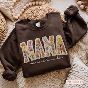 Puede incluir: Una sudadera de cuello redondo en color marrón chocolate oscuro con la palabra MAMA en letras grandes de bloque rellenas de coloridos patrones florales bordados. Debajo, los nombres anna, noha y alana en cursiva blanca.