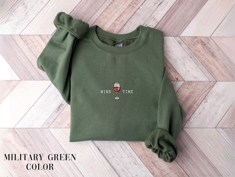 May include: Military green sweatshirt with the words "WINE TIME" and a wine glass graphic. The sweatshirt is made of a soft material and has a crew neck. The sleeves are cuffed at the wrists. The background is a light wood pattern.