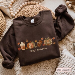 Puede incluir: Una sudadera de cuello redondo en color marrón chocolate oscuro con un estampado gráfico de una taza de café, una calabaza estampada, una rama de canela y una pila de libros atados con un lazo, junto al texto "cozy season" y hojas de otoño.