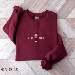 May include: Maroon-colored sweatshirt with the words "WINE TIME" and a wine glass graphic. The sweatshirt is displayed on a light wood-grain surface. The sleeves are folded, and the words "MAROON COLOR" are at the bottom.